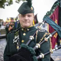A person in a dark green military uniform is standing outside, holding bagpipes. The uniform is adorned with brass buttons and insignia, and they are wearing a matching hat with a feather. There are trees and other people in similar attire visible in the background.