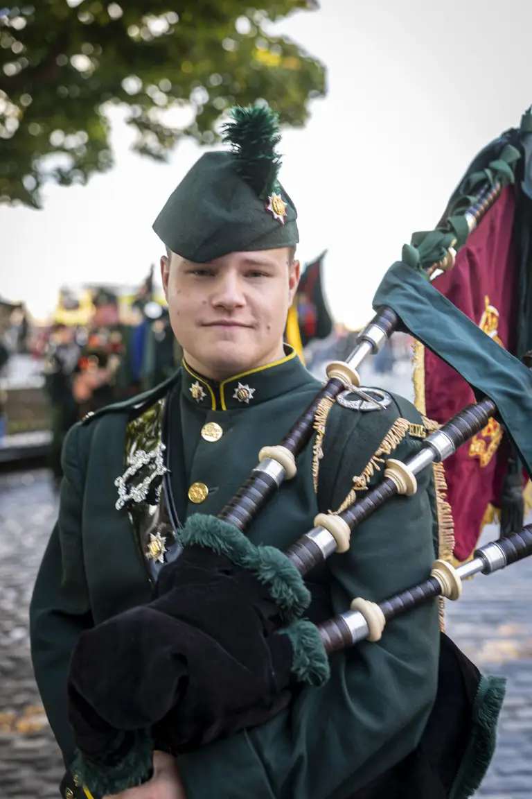 A person in a dark green military uniform is standing outside, holding bagpipes. The uniform is adorned with brass buttons and insignia, and they are wearing a matching hat with a feather. There are trees and other people in similar attire visible in the background.