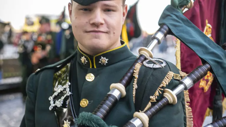 A person in a dark green military uniform is standing outside, holding bagpipes. The uniform is adorned with brass buttons and insignia, and they are wearing a matching hat with a feather. There are trees and other people in similar attire visible in the background.