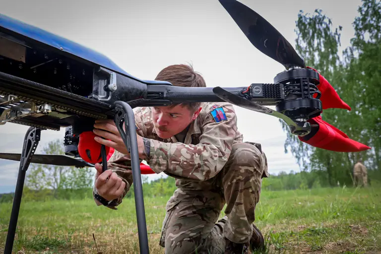 A person in camouflage clothing adjusts a large drone outdoors on a grassy field, with trees in the background. 