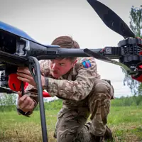 A person in camouflage clothing adjusts a large drone outdoors on a grassy field, with trees in the background.