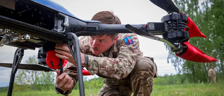 A person in camouflage clothing adjusts a large drone outdoors on a grassy field, with trees in the background.