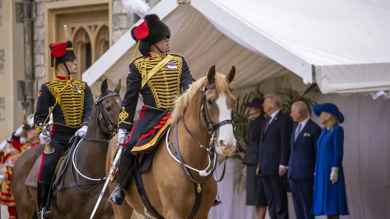 Two ceremonial guards in ornate black and gold uniforms ride horses past a group of formally dressed spectators.