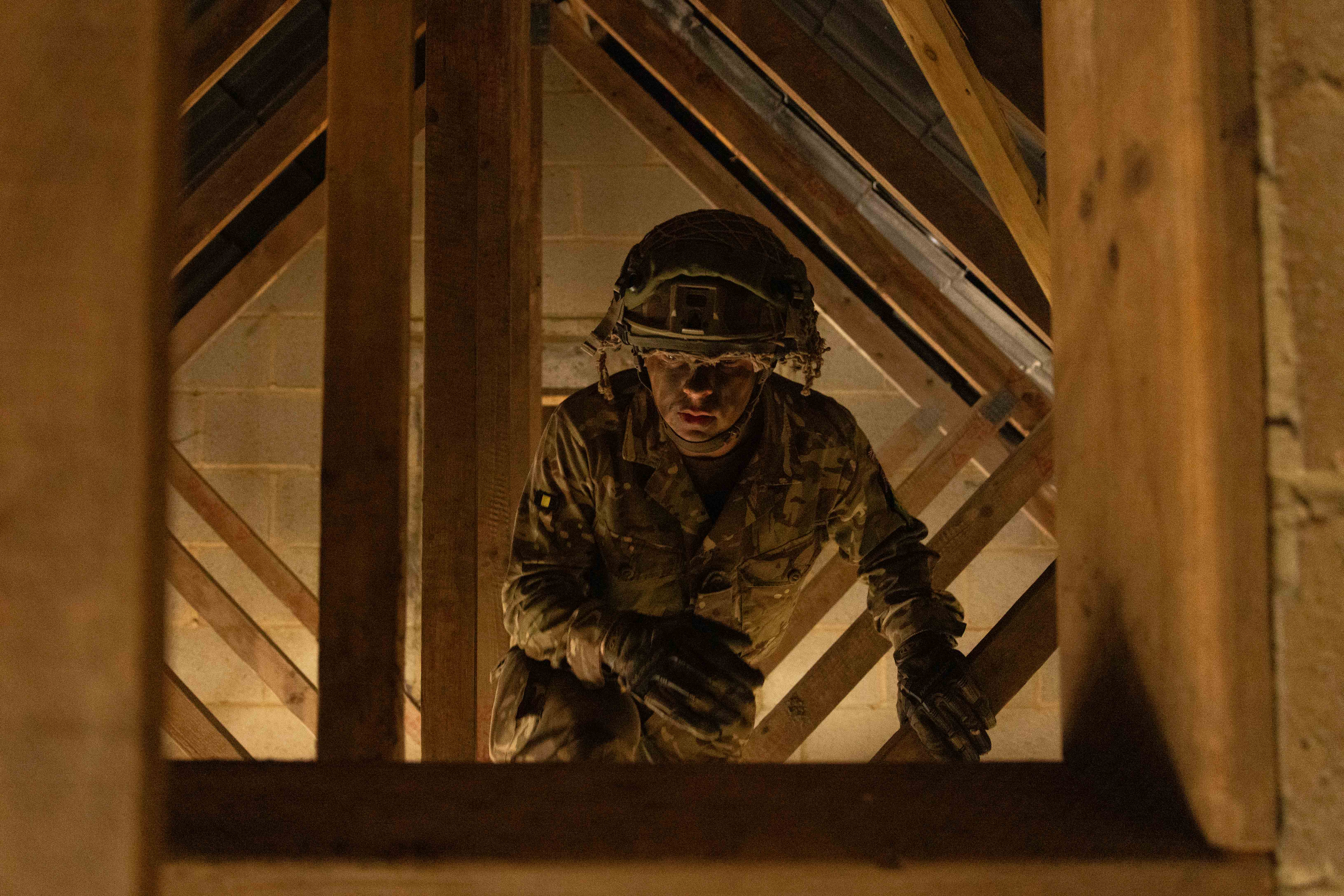 Soldier in camouflage uniform is seen climbing through wooden structure in a loft during assault course training. 