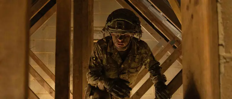 Soldier in camouflage uniform is seen climbing through wooden structure in a loft during assault course training.