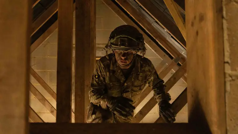 Soldier in camouflage uniform is seen climbing through wooden structure in a loft during assault course training.