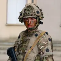 A person in military camouflage gear and helmet stands holding a rifle. Their face is painted with camouflage colours.