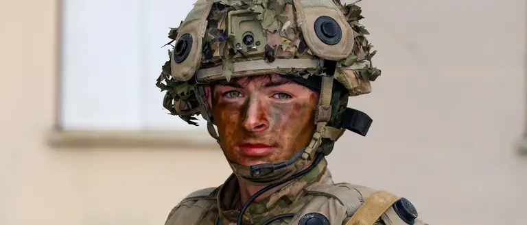 A person in military camouflage gear and helmet stands holding a rifle. Their face is painted with camouflage colours.