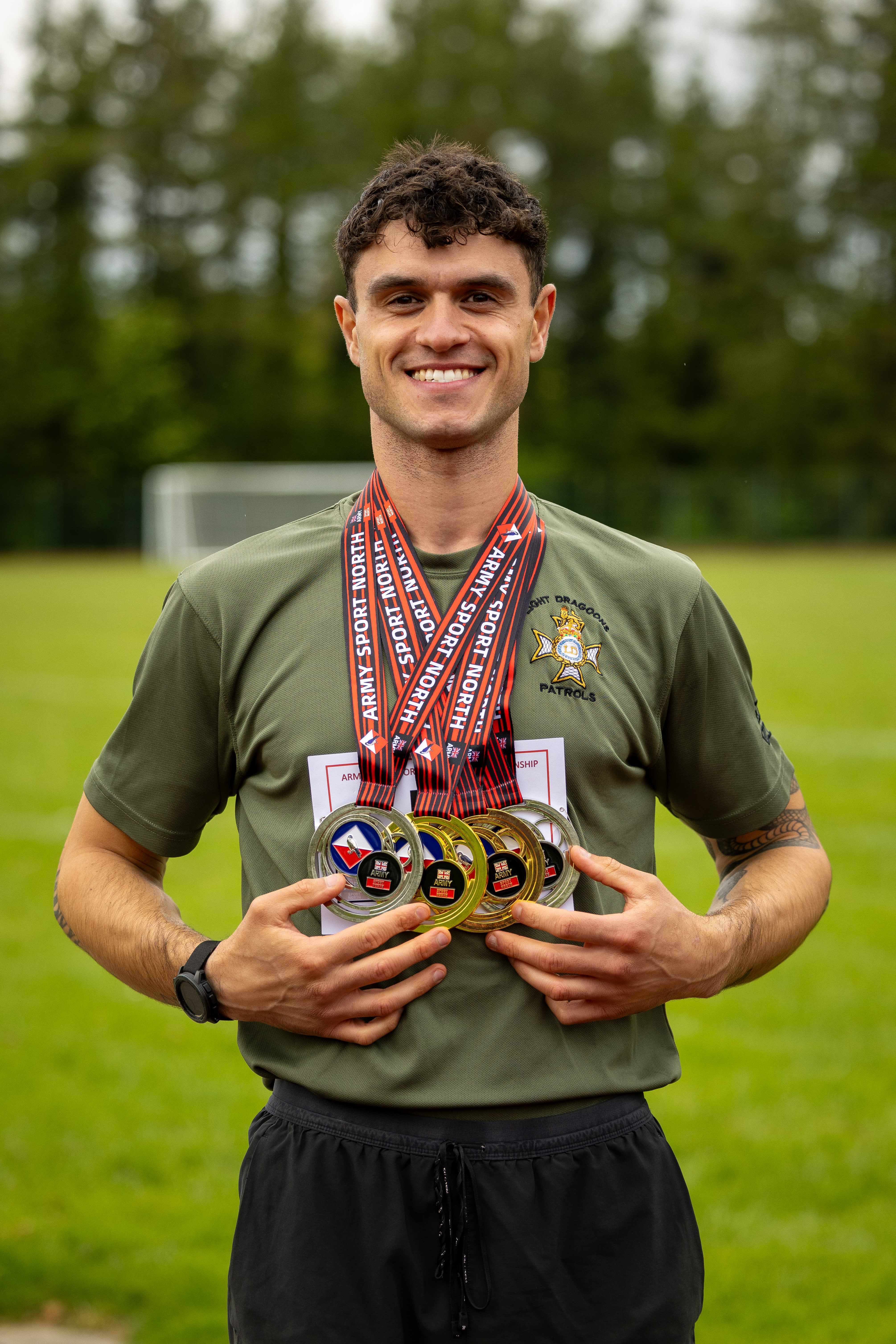 A person standing on a grass field, wearing an olive-green shirt with an emblem, holding multiple medals.