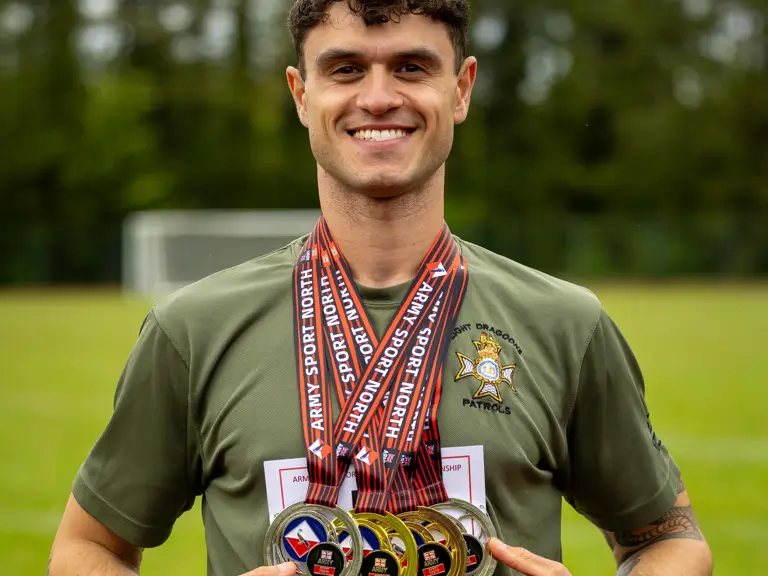 A person standing on a grass field, wearing an olive-green shirt with an emblem, holding multiple medals.