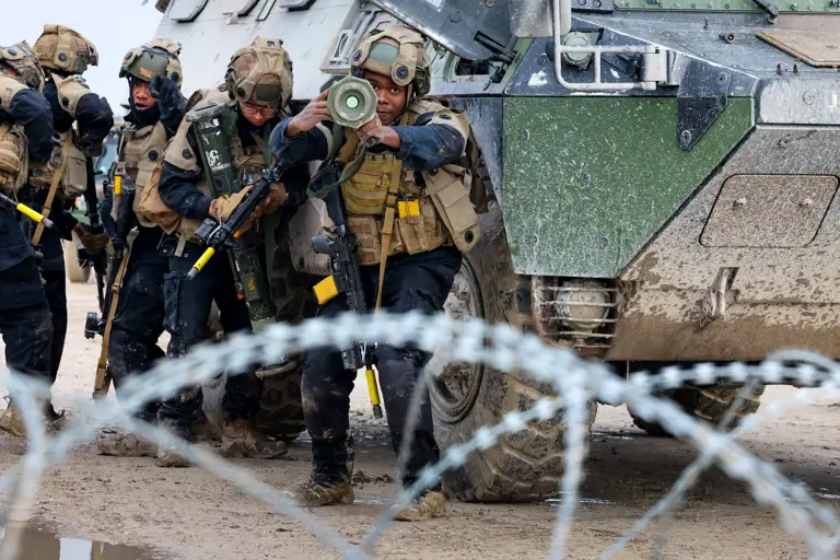 Group of armed soldiers in tactical gear advancing beside an armored military vehicle behind barbed wire.