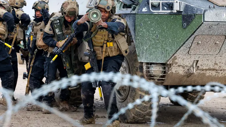 Group of armed soldiers in tactical gear advancing beside an armored military vehicle behind barbed wire.