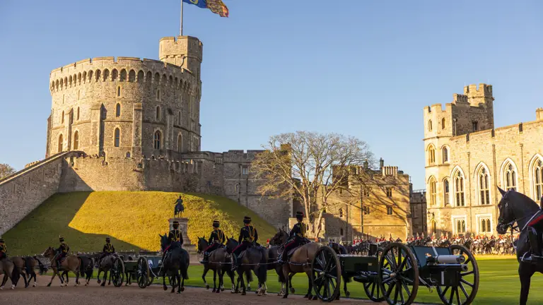 A ceremonial procession with soldiers on horseback and a carriage passes Windsor Castle.