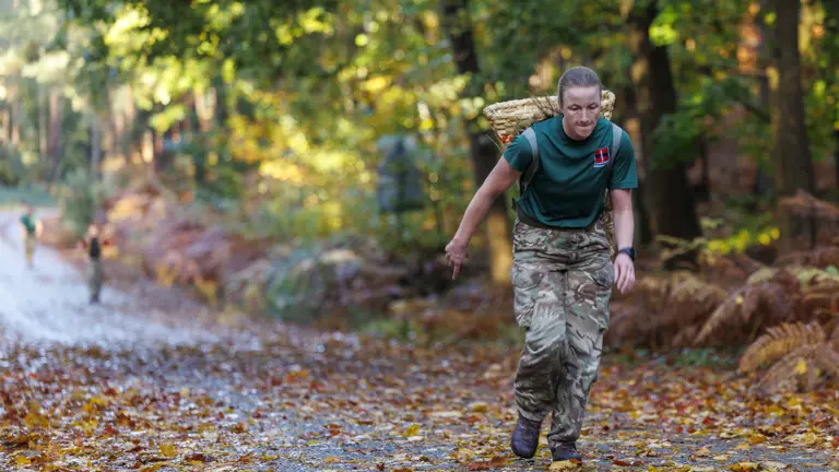 A soldier in camouflage trousers and a green shirt carries a heavy basket on their back while running.