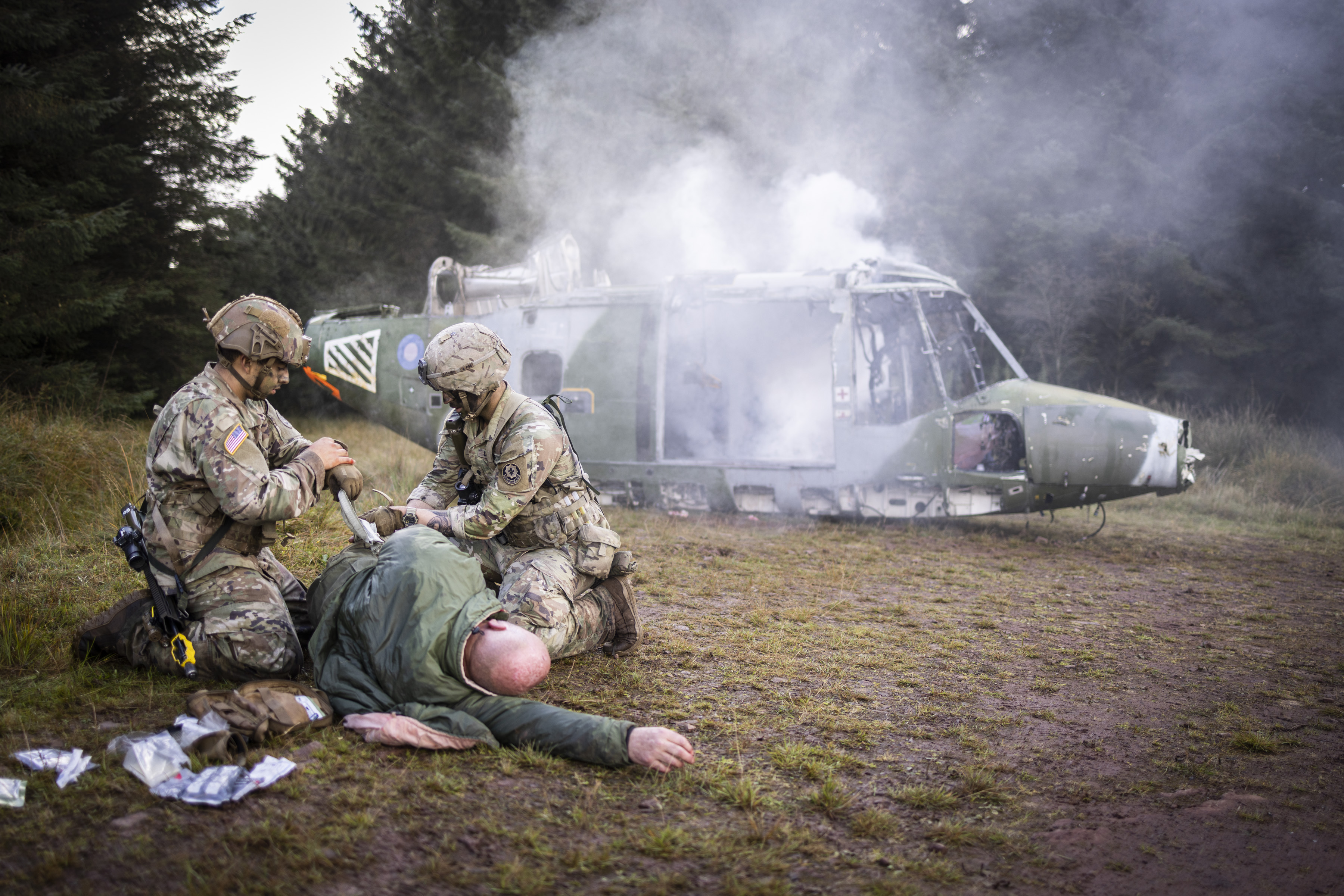 Two soldiers in uniform tend to an injured man in a training exercise, a model helicopter is also present in the background.
