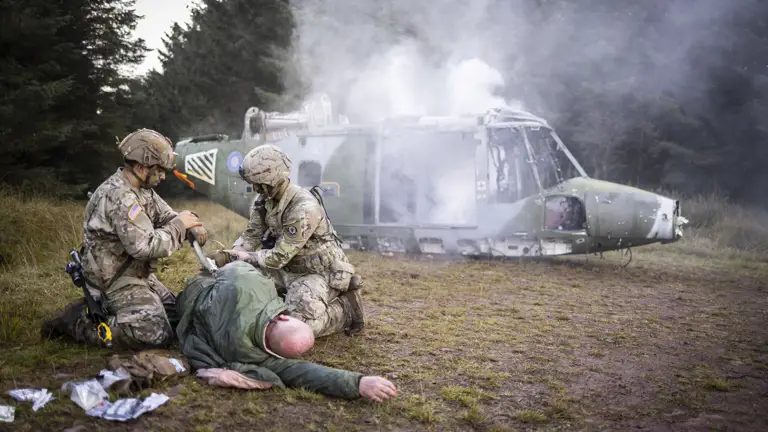 Two soldiers in uniform tend to an injured man in a training exercise, a model helicopter is also present in the background.