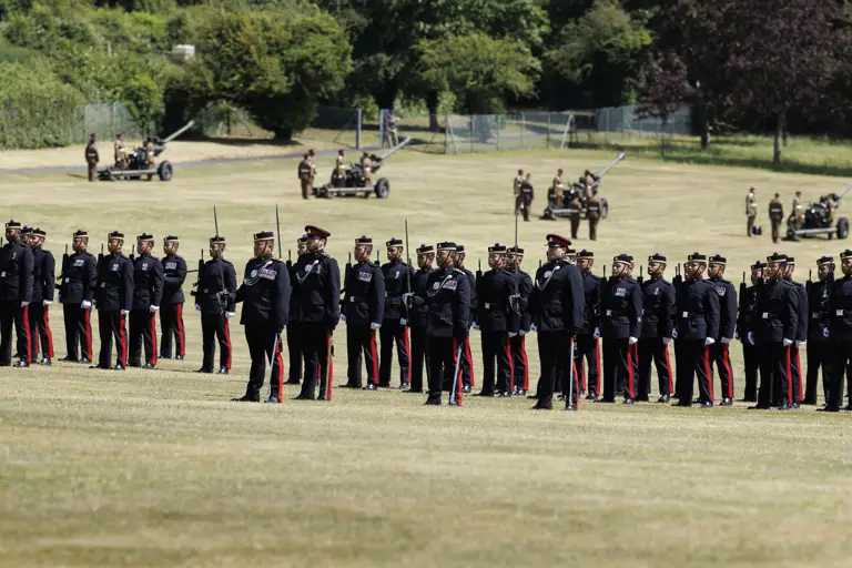 A group of soldiers in formal uniforms stands in formation on a grassy field. 