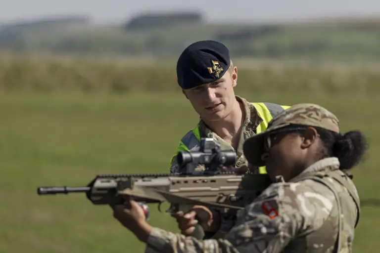 A person in a military uniform assists another holding a rifle, outside on a grassy field. The helper wears a beret.