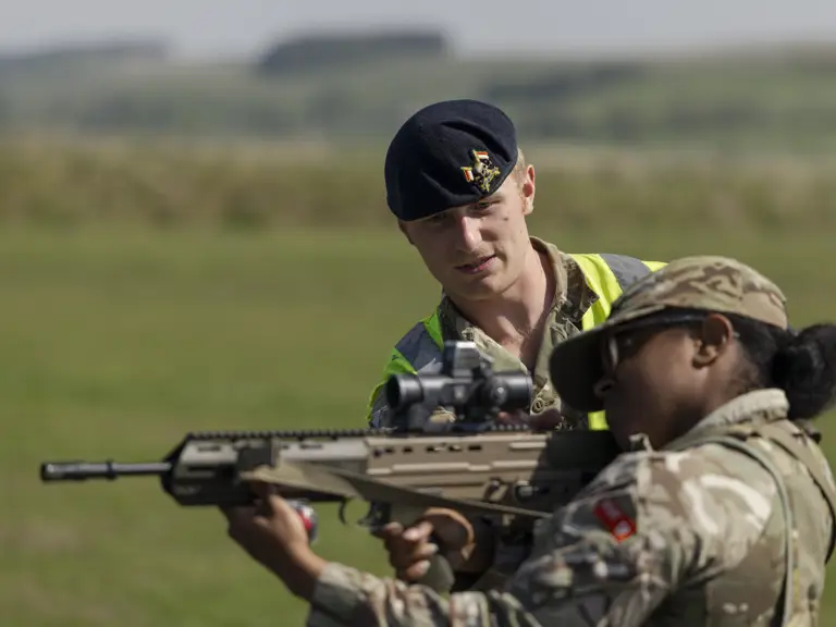 A person in a military uniform assists another holding a rifle, outside on a grassy field. The helper wears a beret.