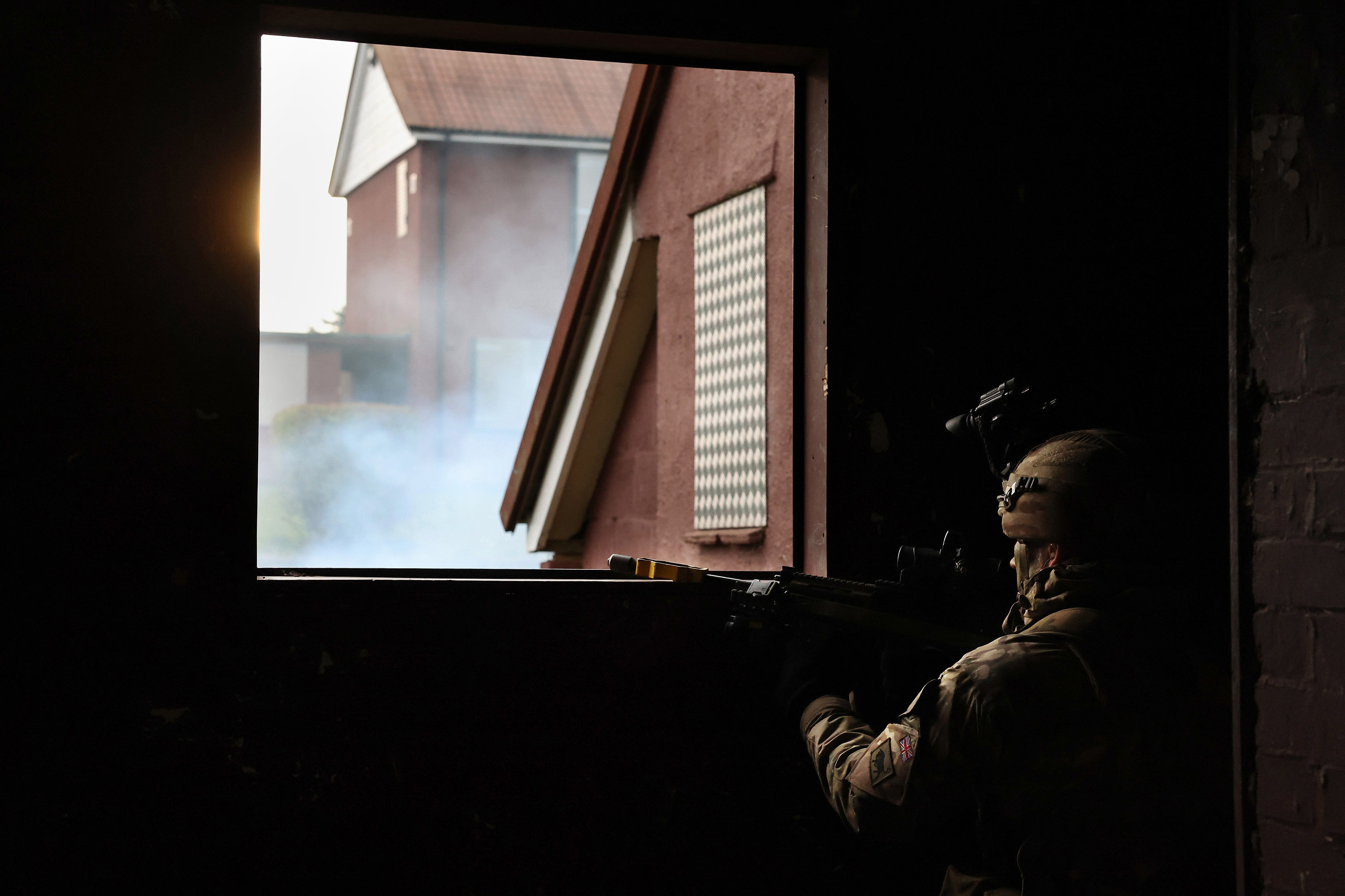 A soldier kneels in darkness next to a window to take aim.