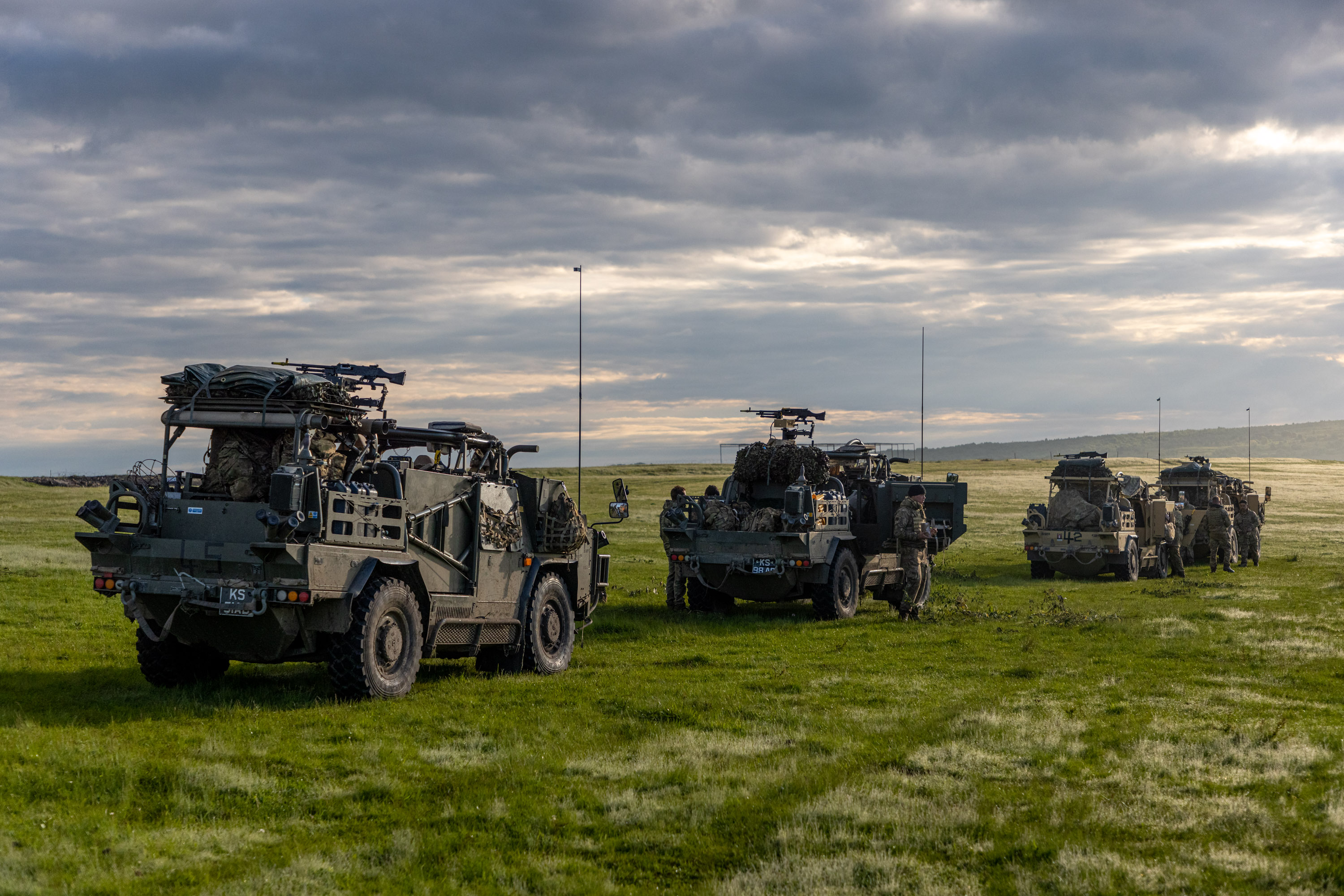 Four Jackal vehicles are driving in convoy through am empty green field with vast space around them. Soldiers are shown walking around the vehicles and the military kit is in the vehicles.