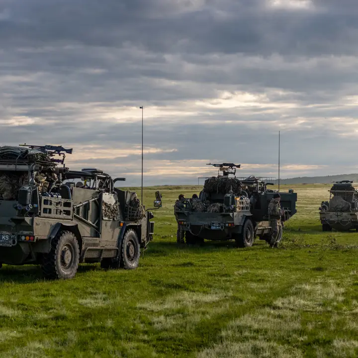 Four Jackal vehicles are driving in convoy through am empty green field with vast space around them. Soldiers are shown walking around the vehicles and the military kit is in the vehicles.