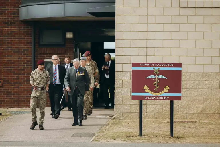 His Royal Highness the Duke of Gloucester walks with soldiers. To their left is a maroon sign which reads