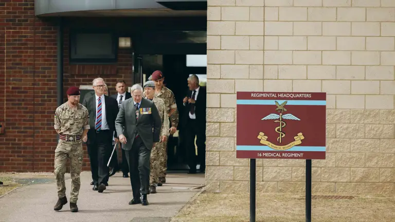 His Royal Highness the Duke of Gloucester walks with soldiers. To their left is a maroon sign which reads