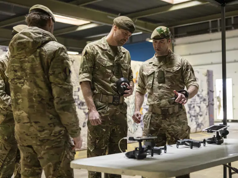 A group of people in military camouflage uniforms are gathered around a table examining drones.