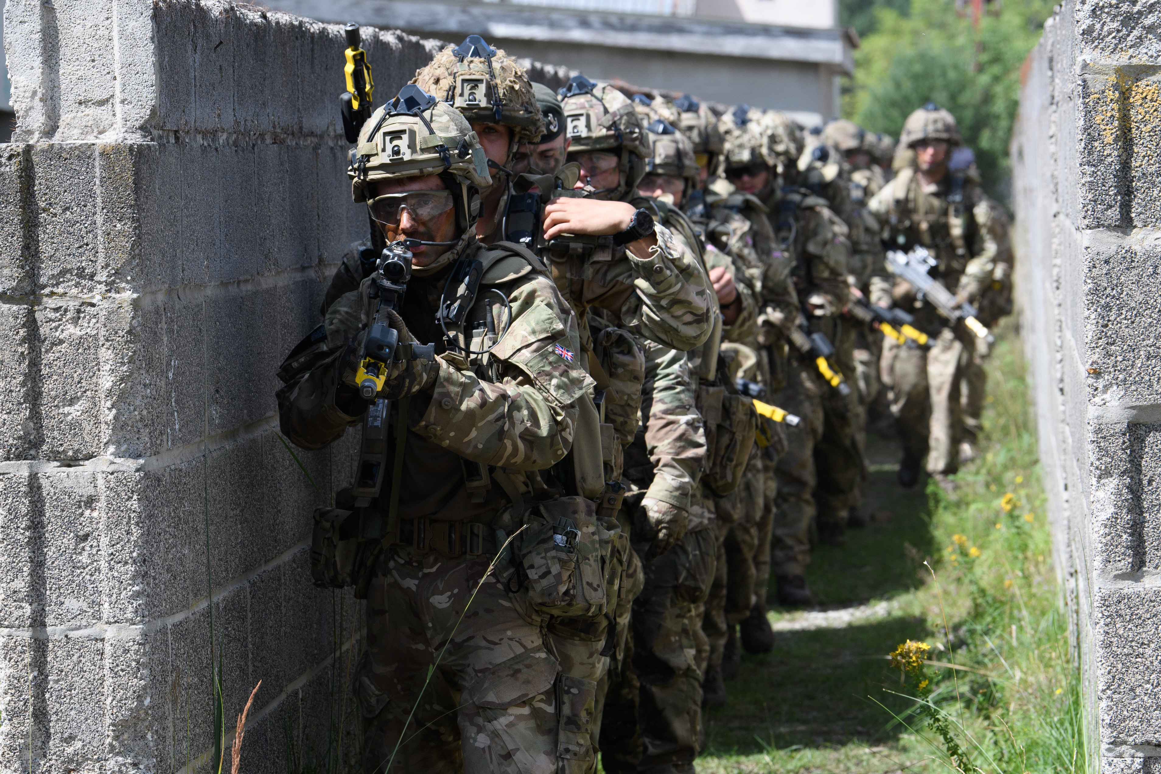 A group of armed soldiers in camouflage uniforms and helmets move along a narrow, walled pathway in a tactical formation. They are equipped with rifles and gear, appearing focused and coordinated. The scene suggests a military exercise or operation in an urban environment.