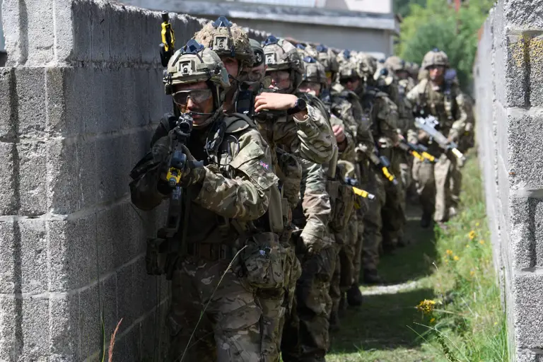 A group of armed soldiers in camouflage uniforms and helmets move along a narrow, walled pathway in a tactical formation. They are equipped with rifles and gear, appearing focused and coordinated. The scene suggests a military exercise or operation in an urban environment.