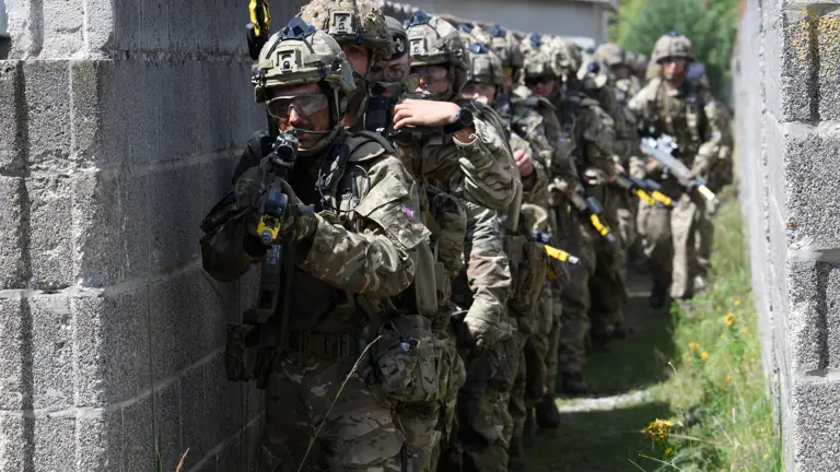 A group of armed soldiers in camouflage uniforms and helmets move along a narrow, walled pathway in a tactical formation. They are equipped with rifles and gear, appearing focused and coordinated. The scene suggests a military exercise or operation in an urban environment.