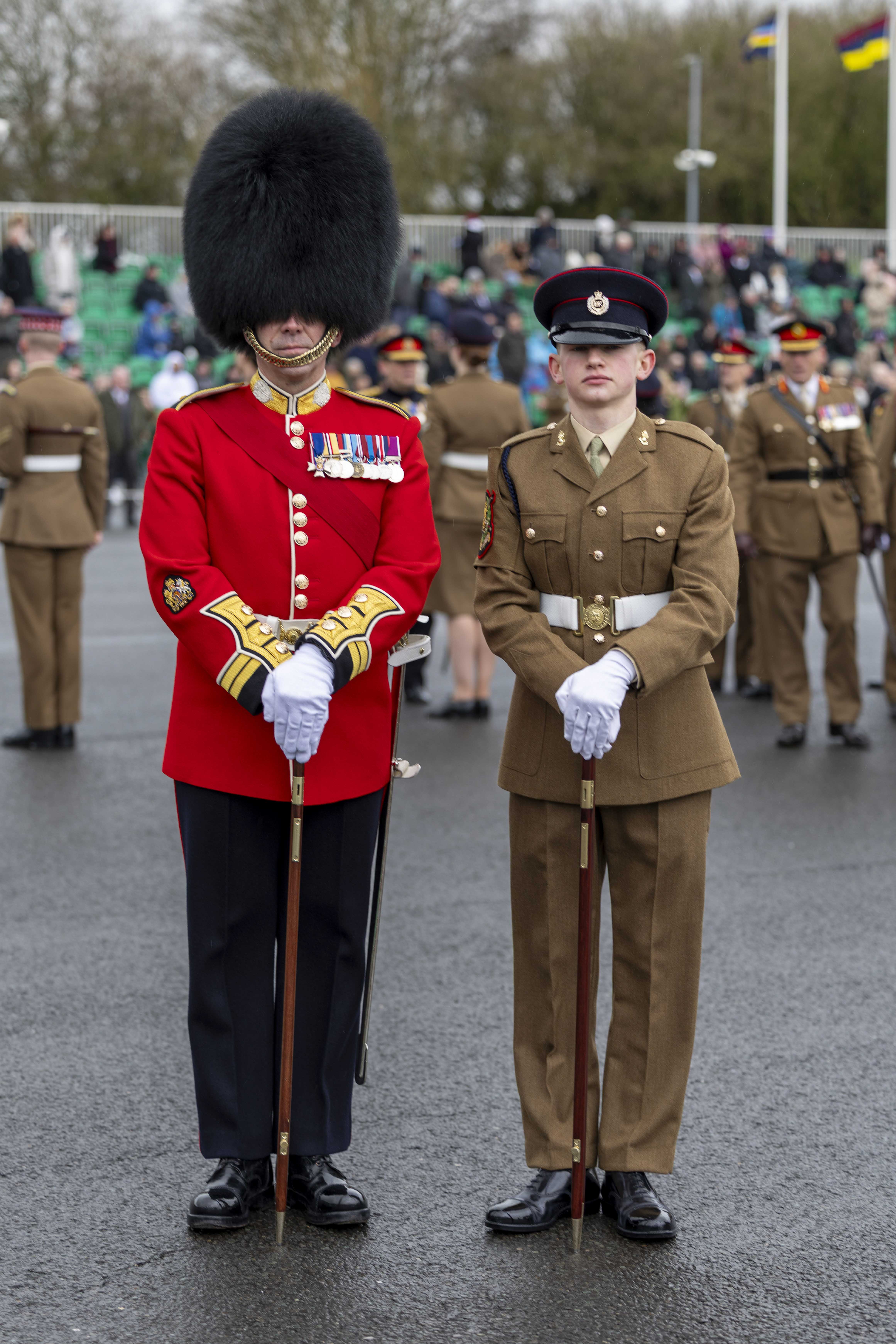 Two soldiers in ceremonial uniforms stand at attention on a parade ground. The one on the left wears a red coat and bearskin hat, and the other is in brown.