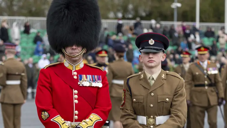 Two soldiers in ceremonial uniforms stand at attention on a parade ground. The one on the left wears a red coat and bearskin hat, and the other is in brown.