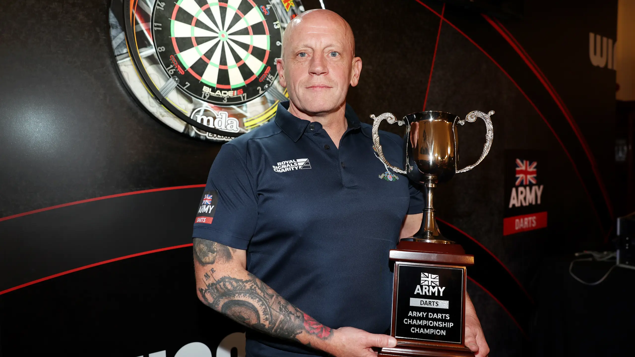 Person holding the Army Darts Championship trophy in front of a dartboard and branded backdrop.