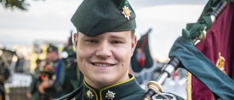 A person in a dark green military uniform is standing outside, holding bagpipes. The uniform is adorned with brass buttons and insignia, and they are wearing a matching hat with a feather. There are trees and other people in similar attire visible in the background.