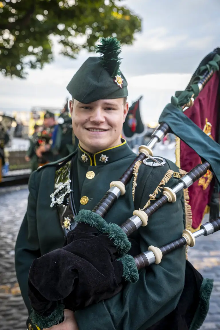 A person in a dark green military uniform is standing outside, holding bagpipes. The uniform is adorned with brass buttons and insignia, and they are wearing a matching hat with a feather. There are trees and other people in similar attire visible in the background.