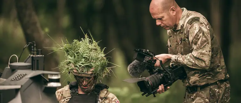 An Army Photographer films a soldier in a wooded area with a modern unmanned vehicle. The photographer is filming on a Canon R5C which has a number of ancillaries such as a larger monitor and microphone. The soldier is wearing his body and helmet which has foliage in to help break up his shape and aid in camouflage.