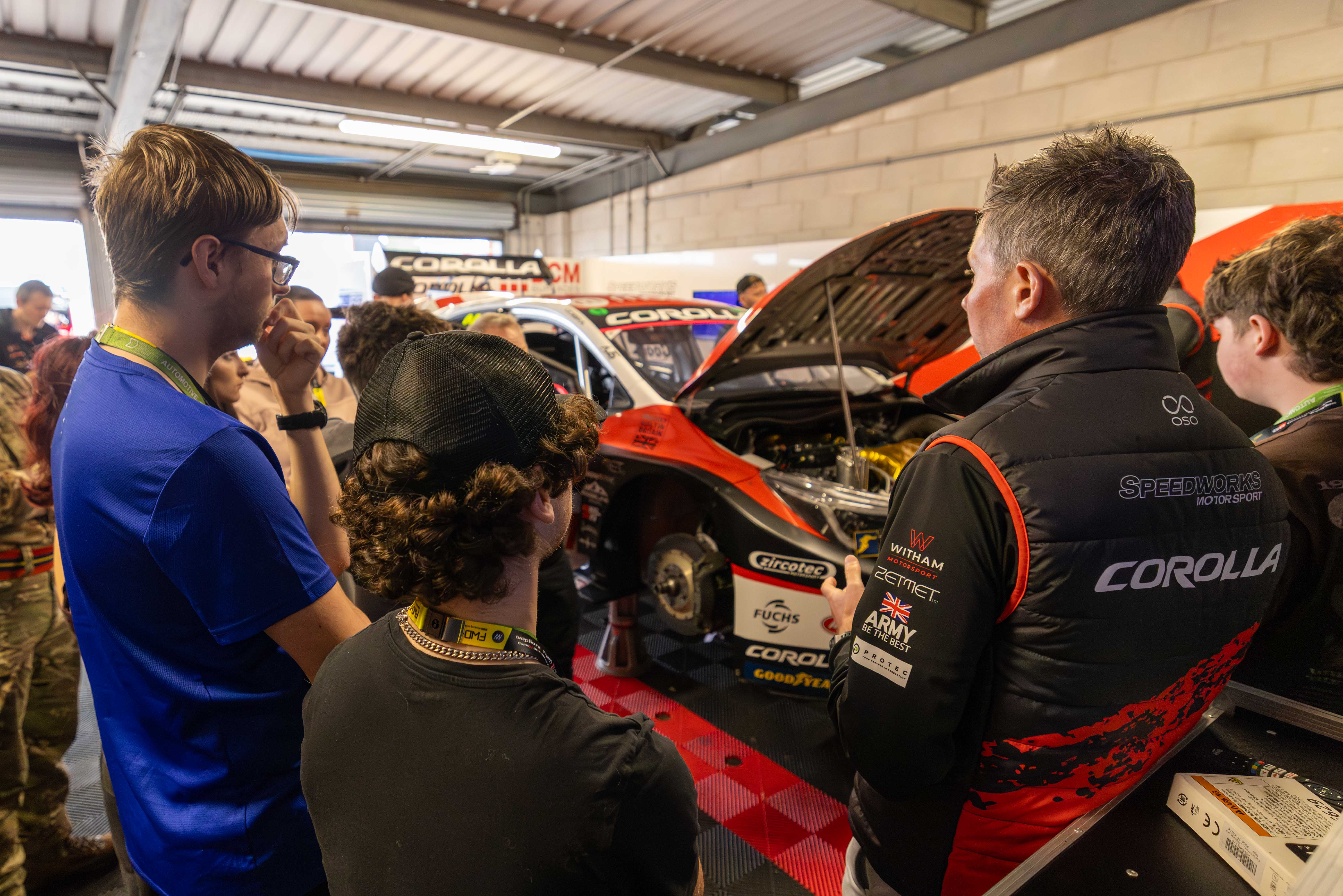 A group of people attentively listen to a person explaining a race car in a garage. The car's hood is open, revealing its engine. The scene conveys interest and focus.
