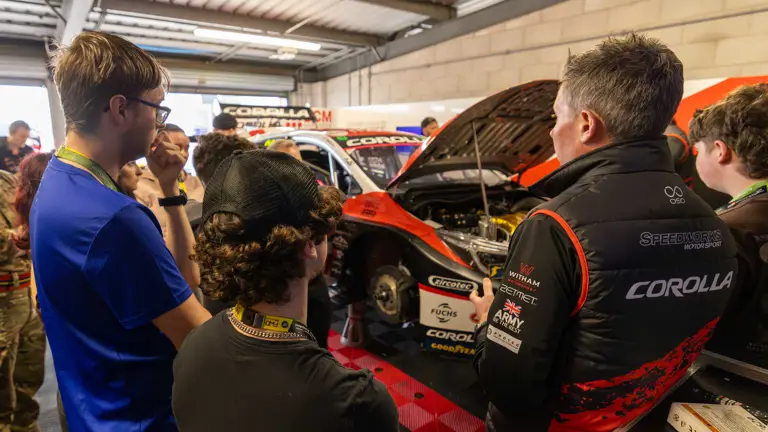 A group of people attentively listen to a person explaining a race car in a garage. The car's hood is open, revealing its engine. The scene conveys interest and focus.