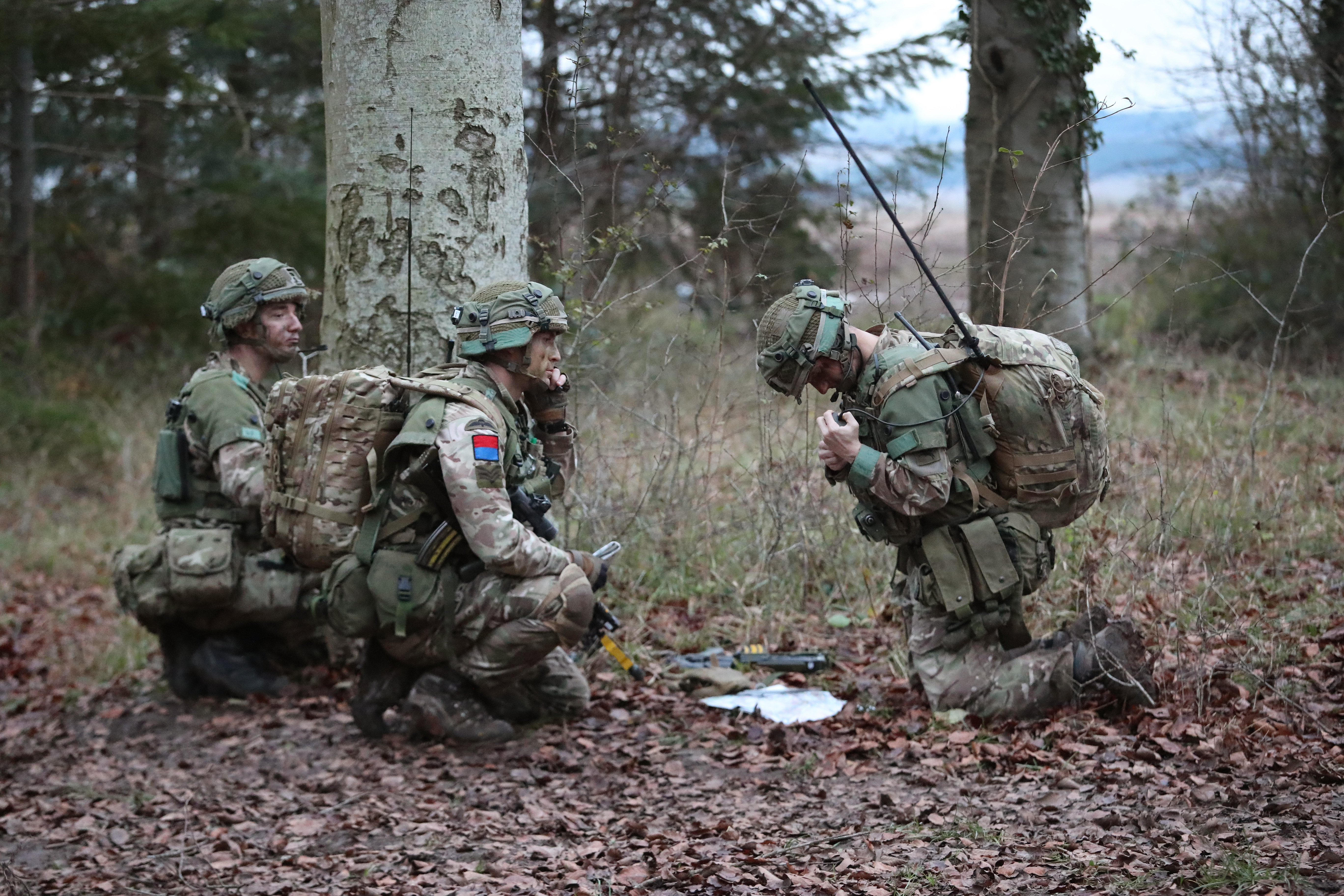 Three soldiers in camouflage gear and backpacks kneel in a forested area, working with communication equipment.