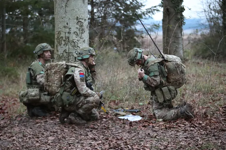 Three soldiers in camouflage gear and backpacks kneel in a forested area, working with communication equipment.