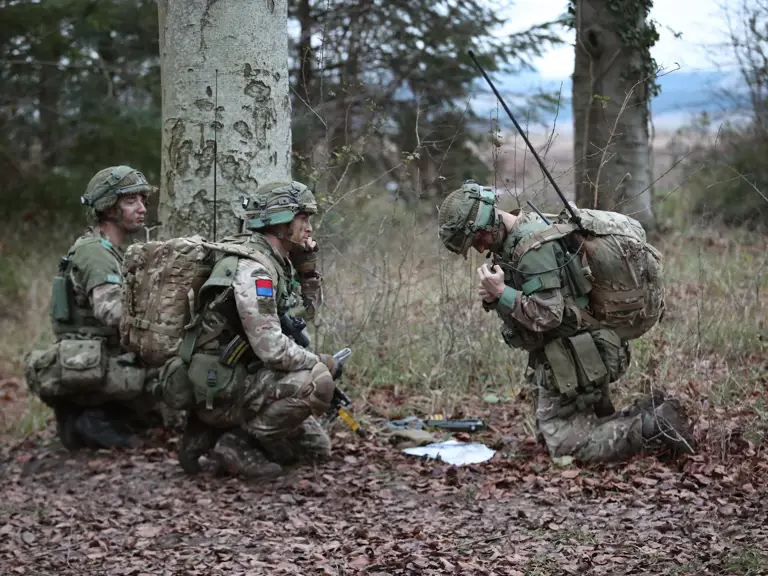 Three soldiers in camouflage gear and backpacks kneel in a forested area, working with communication equipment.