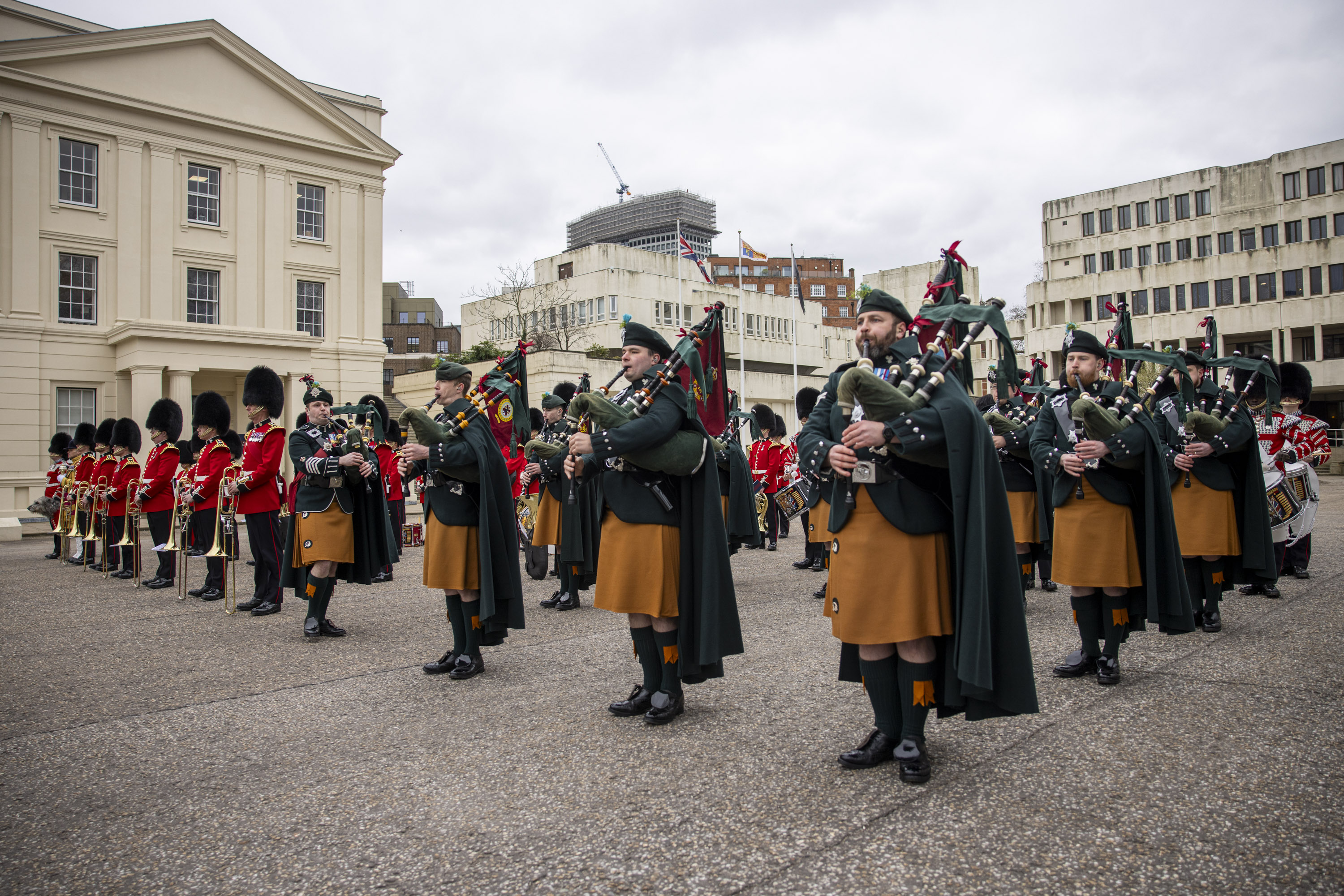 Pipers in green and orange ceremonial attire are pictured next to other soldiers in red tunics and bearskin hats.