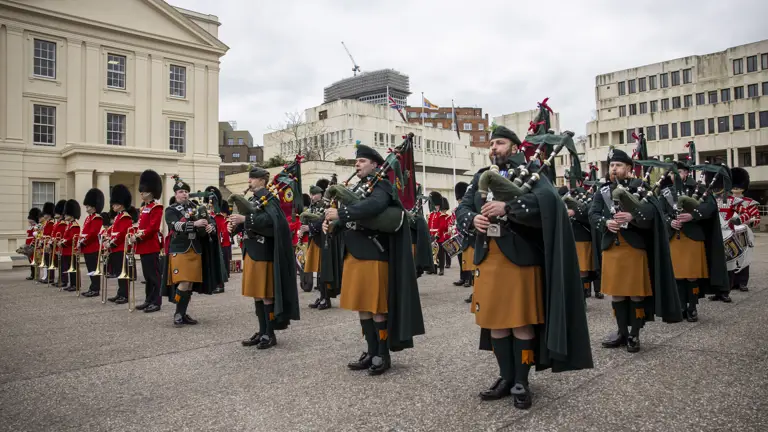 Pipers in green and orange ceremonial attire are pictured next to other soldiers in red tunics and bearskin hats.