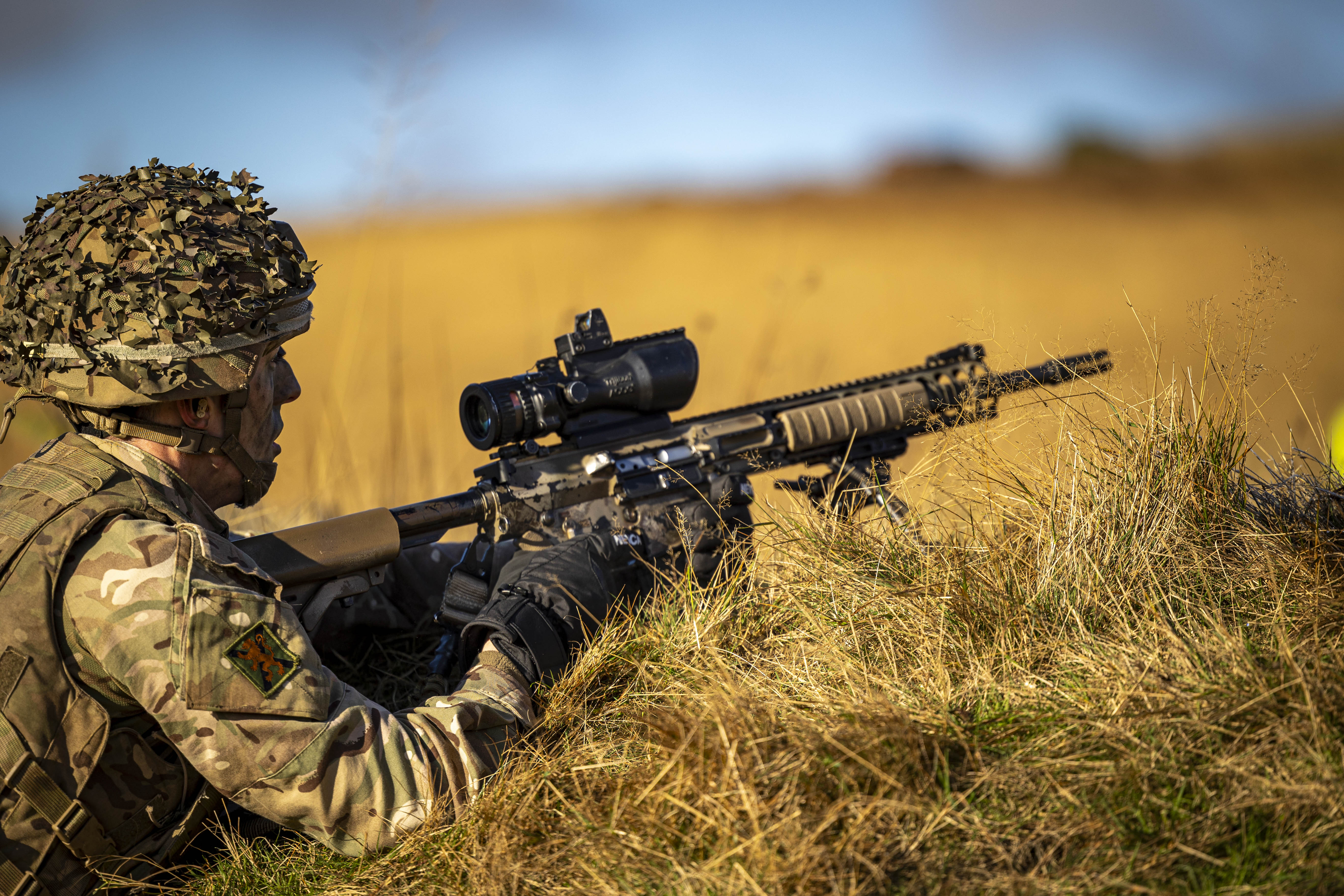 A man in uniform lies prone on the grass with a weapon. 