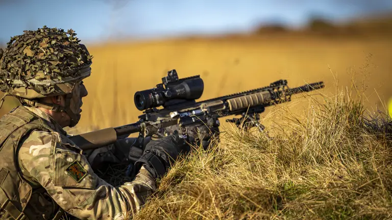 A man in uniform lies prone on the grass with a weapon.