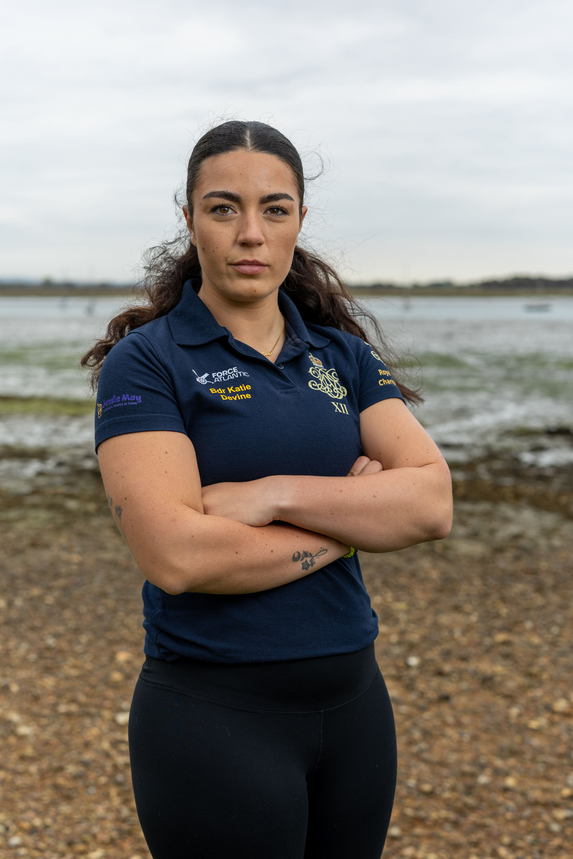 A female Army soldier, wearing dark blue shirt with an Army cap badge on it, is standing on a shoreline with an estuary in the background.