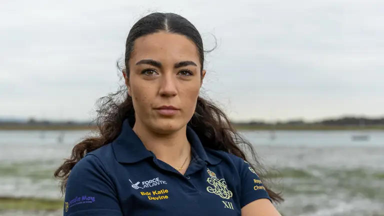 A female Army soldier, wearing dark blue shirt with an Army cap badge on it, is standing on a shoreline with an estuary in the background.