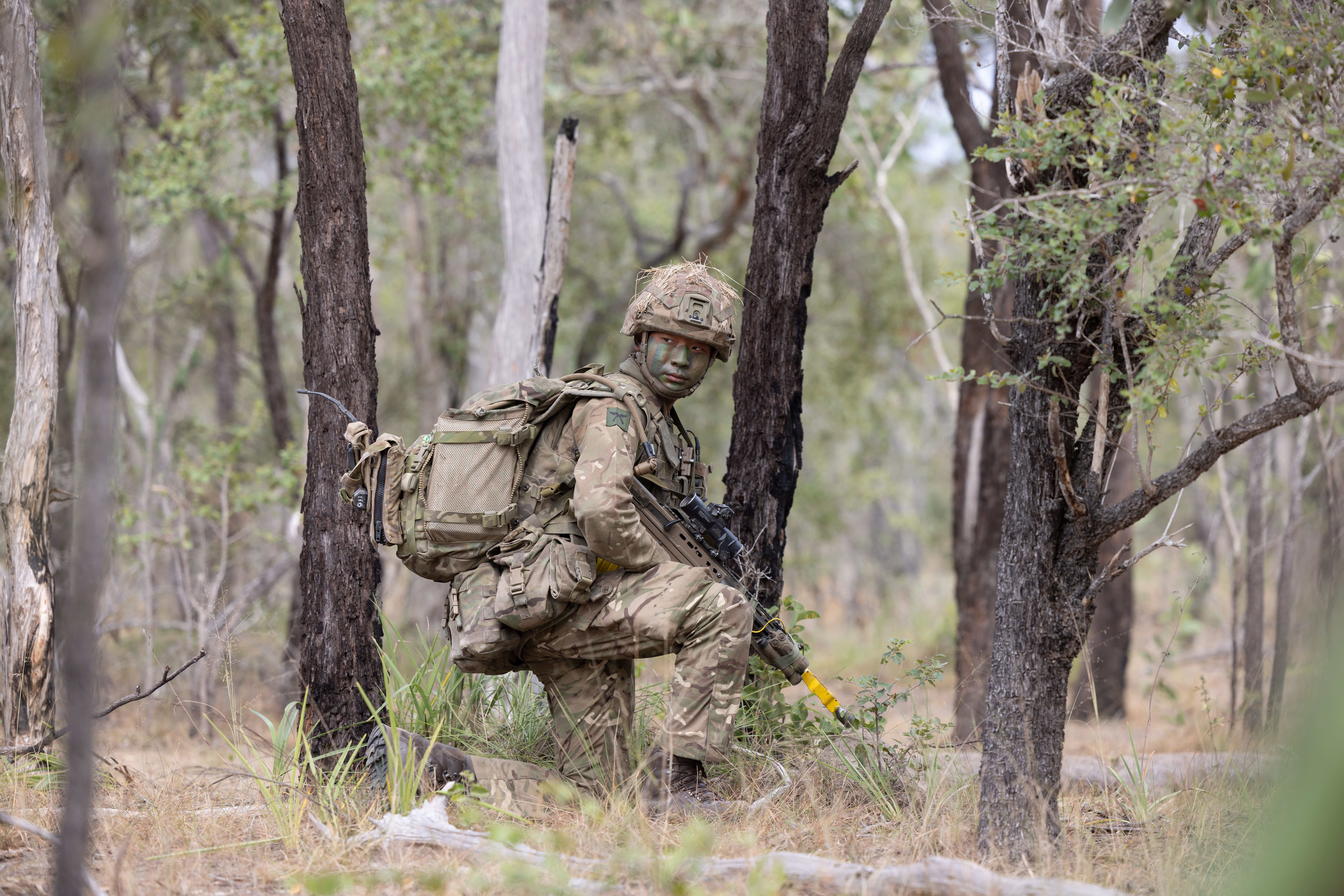 A person in military camouflage, including face paint and gear, is positioned in a forested area, holding a rifle. 